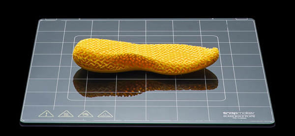 A yellow, 3D-printed shoe insole with a complex lattice structure sits on a glass build plate, showing a popular use for flexible TPU filament.