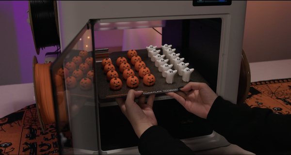 Hands remove a 3D printer build plate covered in freshly printed multicolor pumpkin and ghost game pieces for the Halloween balance game.