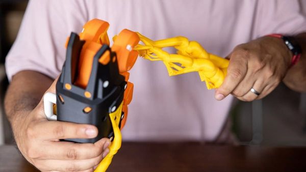 A person's hands carefully separating a bright yellow 3D printed support structure from a black and orange mechanical part.