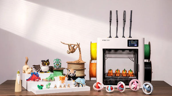 A Snapmaker 3D printer sits on a desk surrounded by colorful 3D-printed models, including articulated dinosaurs, a hanging sloth, and festive pumpkins.