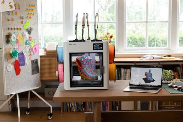 A desktop workspace featuring a Snapmaker 3D printer holding a multi-color shoe print next to a laptop displaying the slicer software.