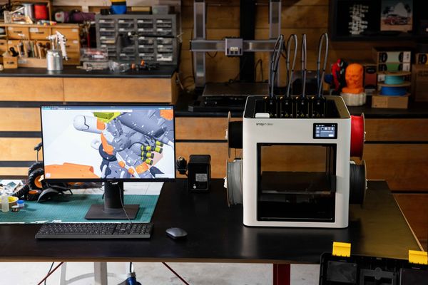 A Snapmaker U1 3D printer sits on a workbench next to a computer monitor displaying pre-processing slicing software, workflow from digital mesh translation to physical hardware execution.
