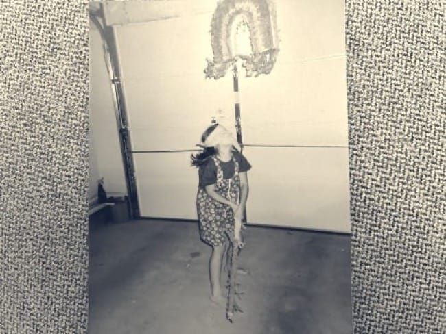 An older child, blindfolded, preparing to hit a piñata hanging in a garage. She is wearing a purple shirt and purple and green flower overalls.