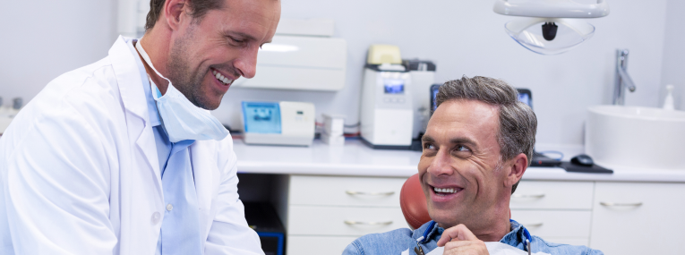 Ein auf dem Behandlungsstuhl sitzender Patient und sein Zahnarzt lächeln sich an. 