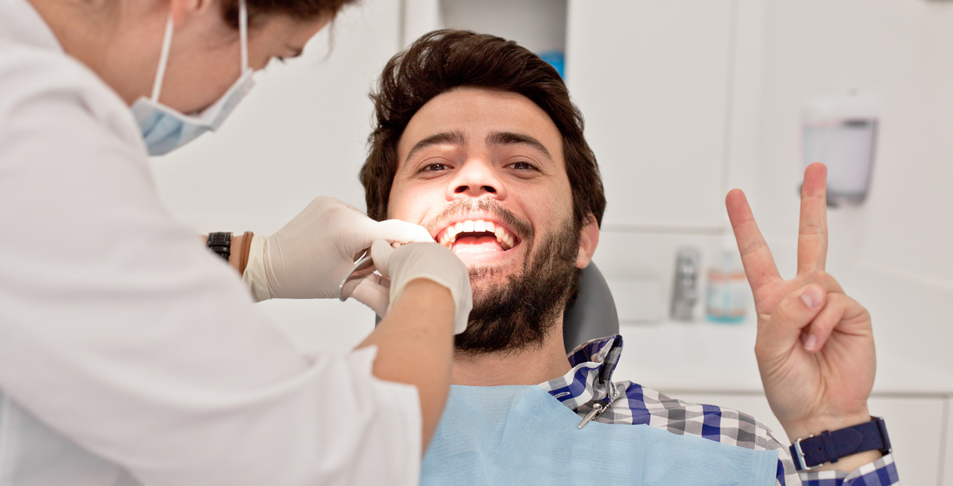 Ein lächelnder Patient wird von einer Zahnärztin behandelt und zeigt dabei ein Peace-Zeichen in die Kamera.