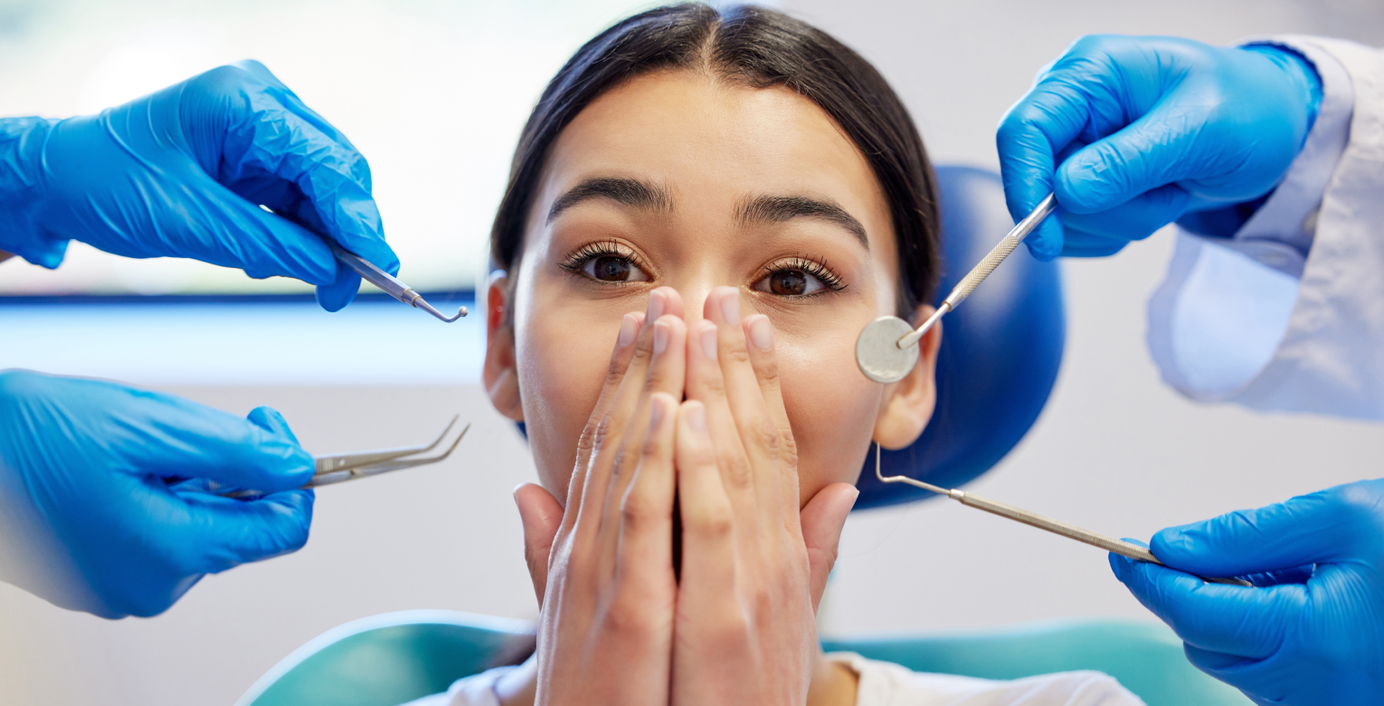 Eine Angstpatientin hält sich vor Schock die Hände vor den Mund, während beim Zahnzart mehrere Behandlungsinstrumente vor sie gehalten werden.