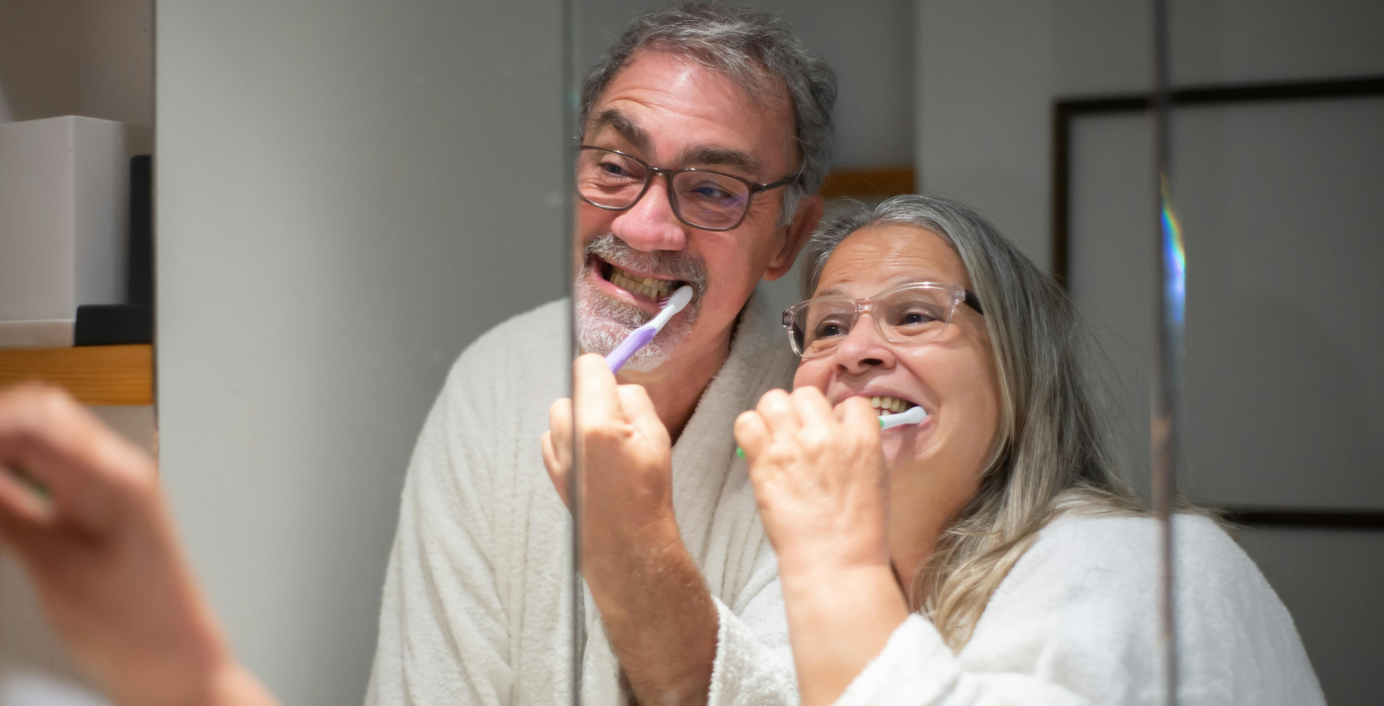 Ein Seniorenpaar schaut beim Zähneputzen in den Spiegel.