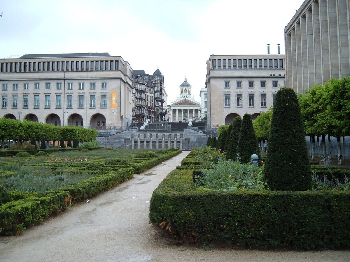 Le jardin du Mont des Arts à Bruxelles, avec ses haies taillées et une allée centrale entre deux bâtiments en pierre claire, et au fond une église surmontée d'une coupole.