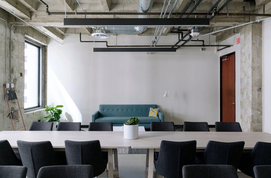 Espacio de coworking luminoso con una mesa larga de madera, sillas grises y un sofá frente a una pared blanca.