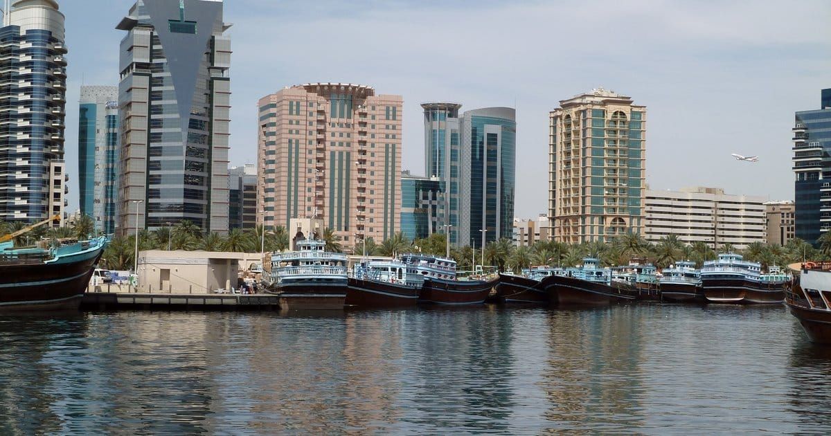 Wooden dhows moored on Dubai Creek in front of Deira towers, with a plane landing overhead.