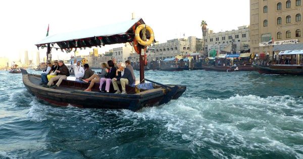 Ein traditionelles Abra-Boot mit Fahrgästen überquert den Dubai Creek, im Hintergrund stehen Gebäude am Ufer von Bur Dubai.