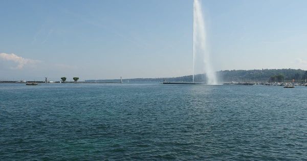 Le lac Léman à Genève sous un ciel bleu, avec le Jet d'eau qui jaillit près d'une jetée et, à gauche, les môles du port avec un petit phare.