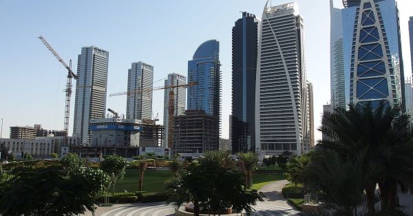 Ein Park mit Palmen und Wegen liegt vor den Hochhäusern von Jumeirah Lakes Towers in Dubai, dem Gebiet der Freezone DMCC, und zwischen den Türmen ragen Baukräne auf.