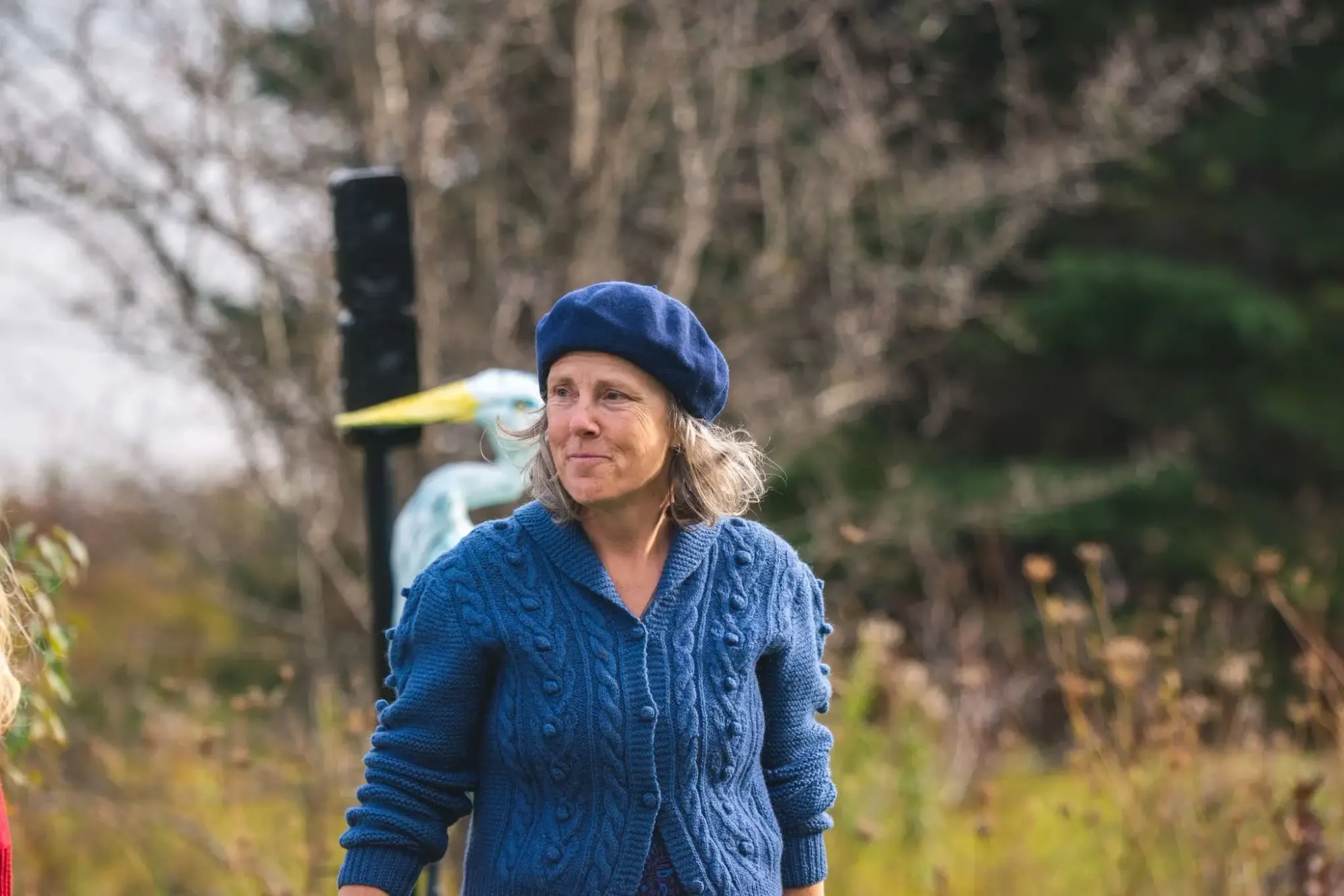 A middle-aged white woman in a blue sweater and beret stands in a field that is also a performance space.