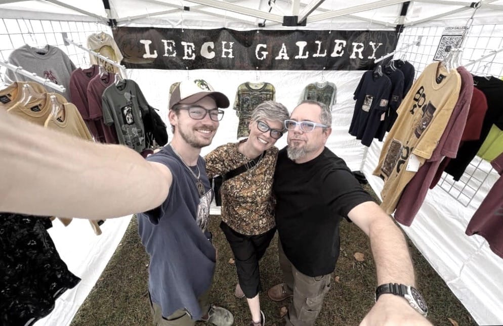 A family of artists smile for a selfie at their booth at an art fair. The banner behind them reads "Leech Gallery."