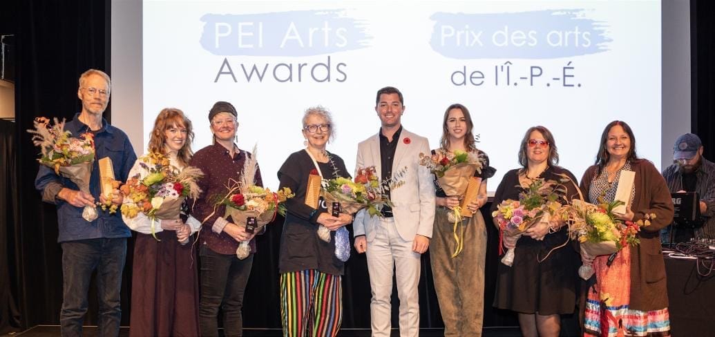 A line of PEI Arts Award recipients of different ages and genders hold their awards and flower bouquets as they pose for a group photo.