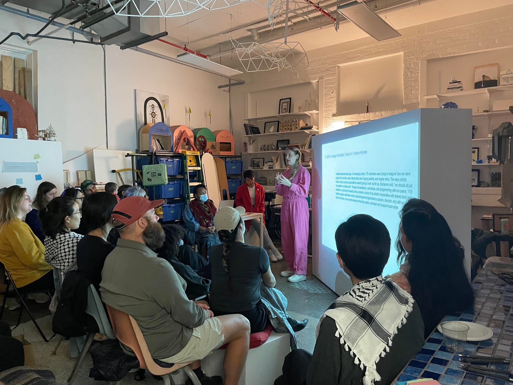 A group of young audience members sit in a white studio space, listening as a blonde woman in a pink jumpsuit speaks in front of a projected slide of text.
