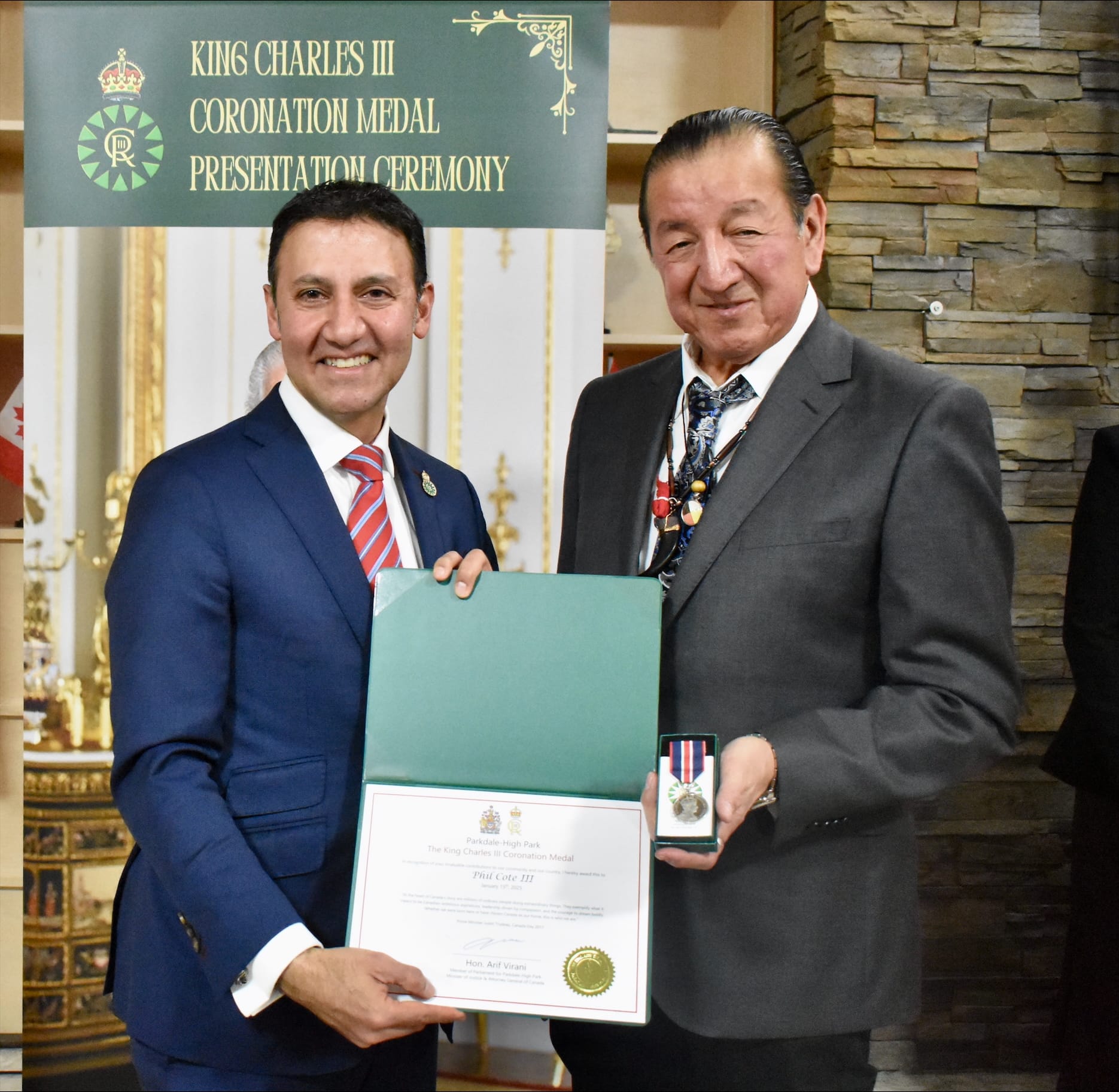 Artist Philip Cote, dressed in a grey suit and dark tie, poses with his Coronation medal and certificate with MP Arif Virani at his side. Both men are smiling.