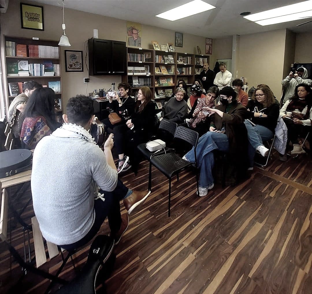 An audience seated in rows in a bookshop, listening to three musicians perform.