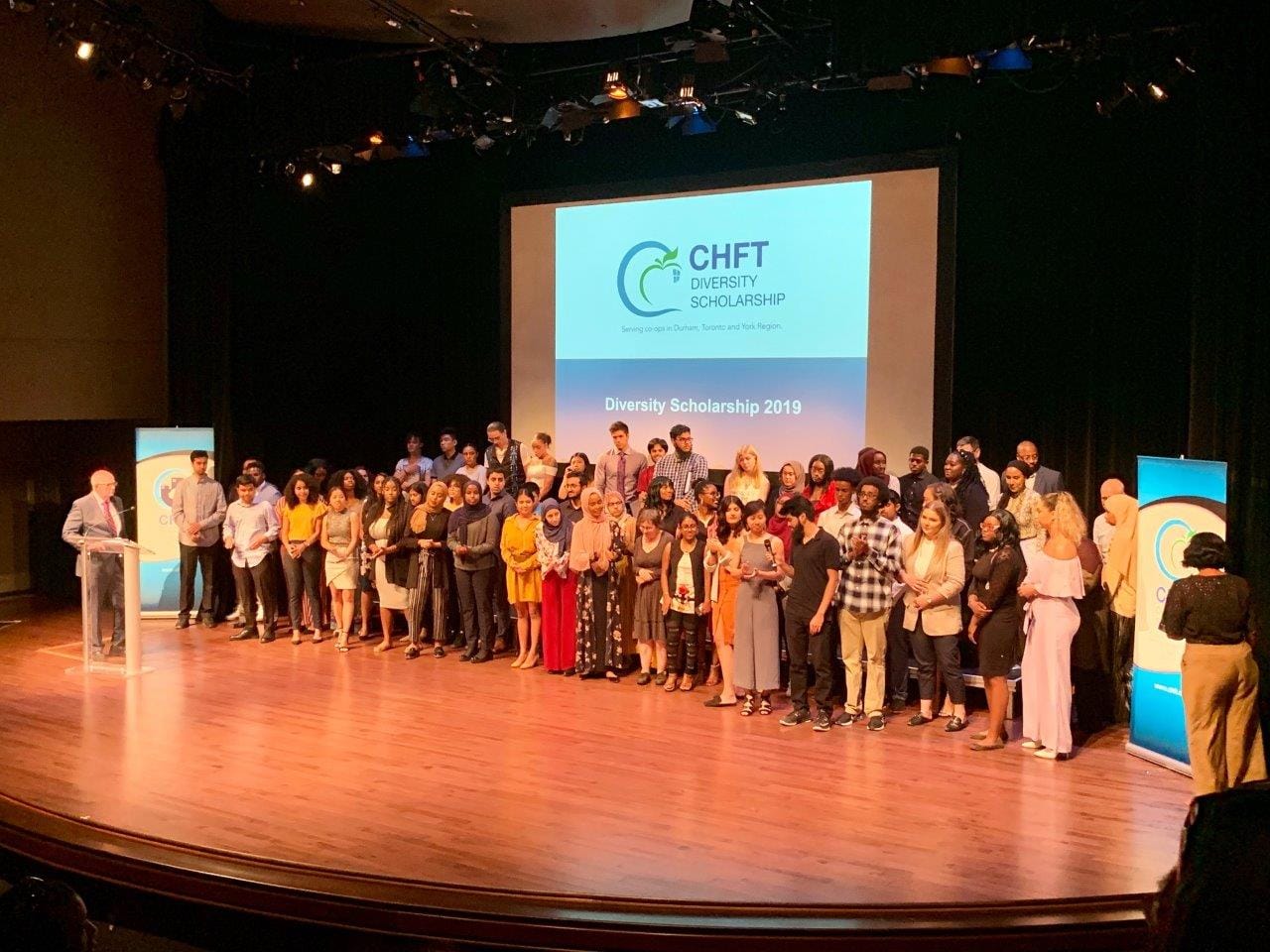 A large crowd of students receiving scholarship awards stands on a stage. Behind them, a slide reading "CHFT Diversity Scholarship" is projected on a screen.