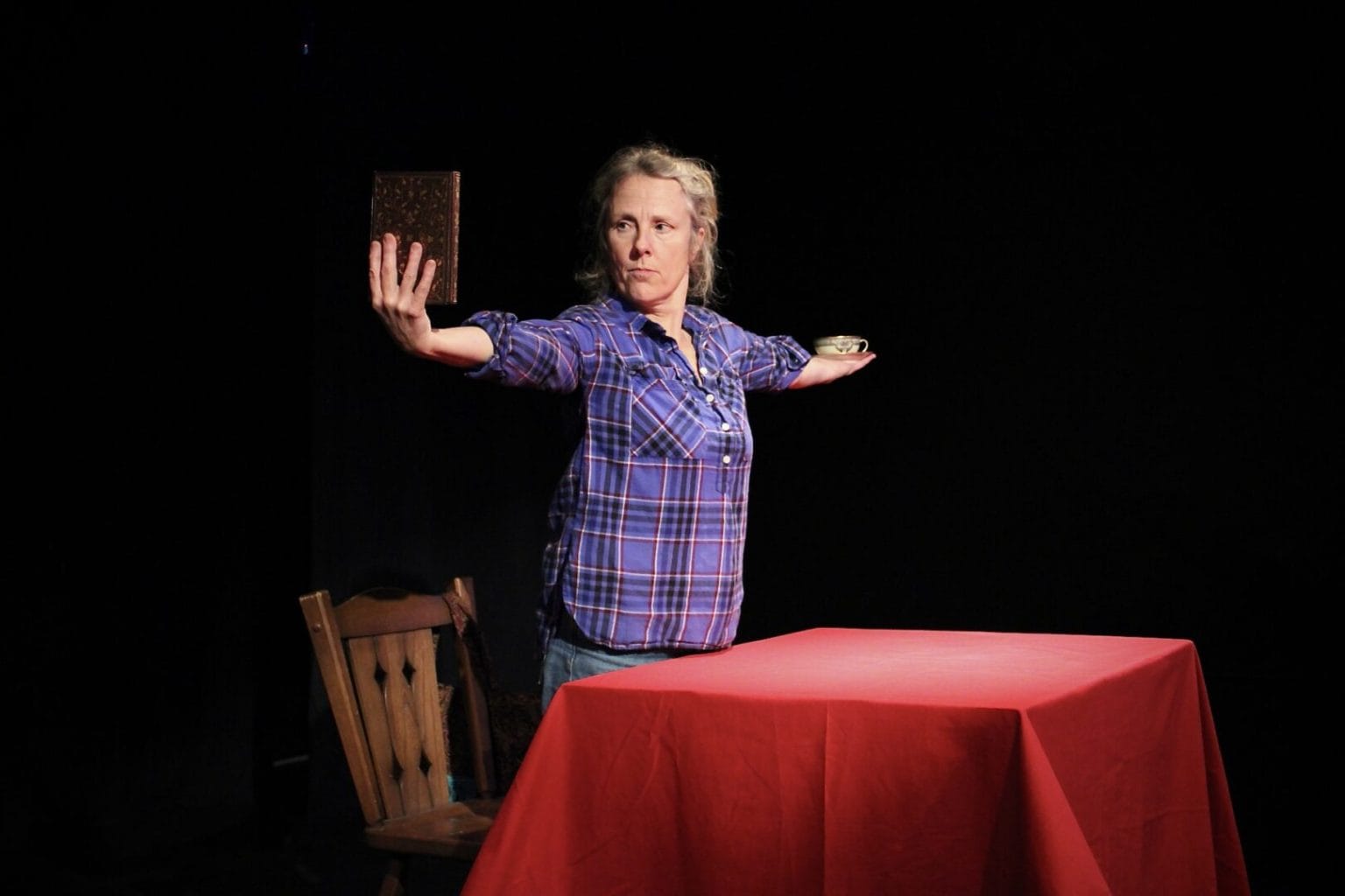 A middle-aged white woman in a plaid shirt stands in front of a table on a darkened stage. Both of her arms are outstretched and she is holding a closed book in her right hand, which she gazes at intently.