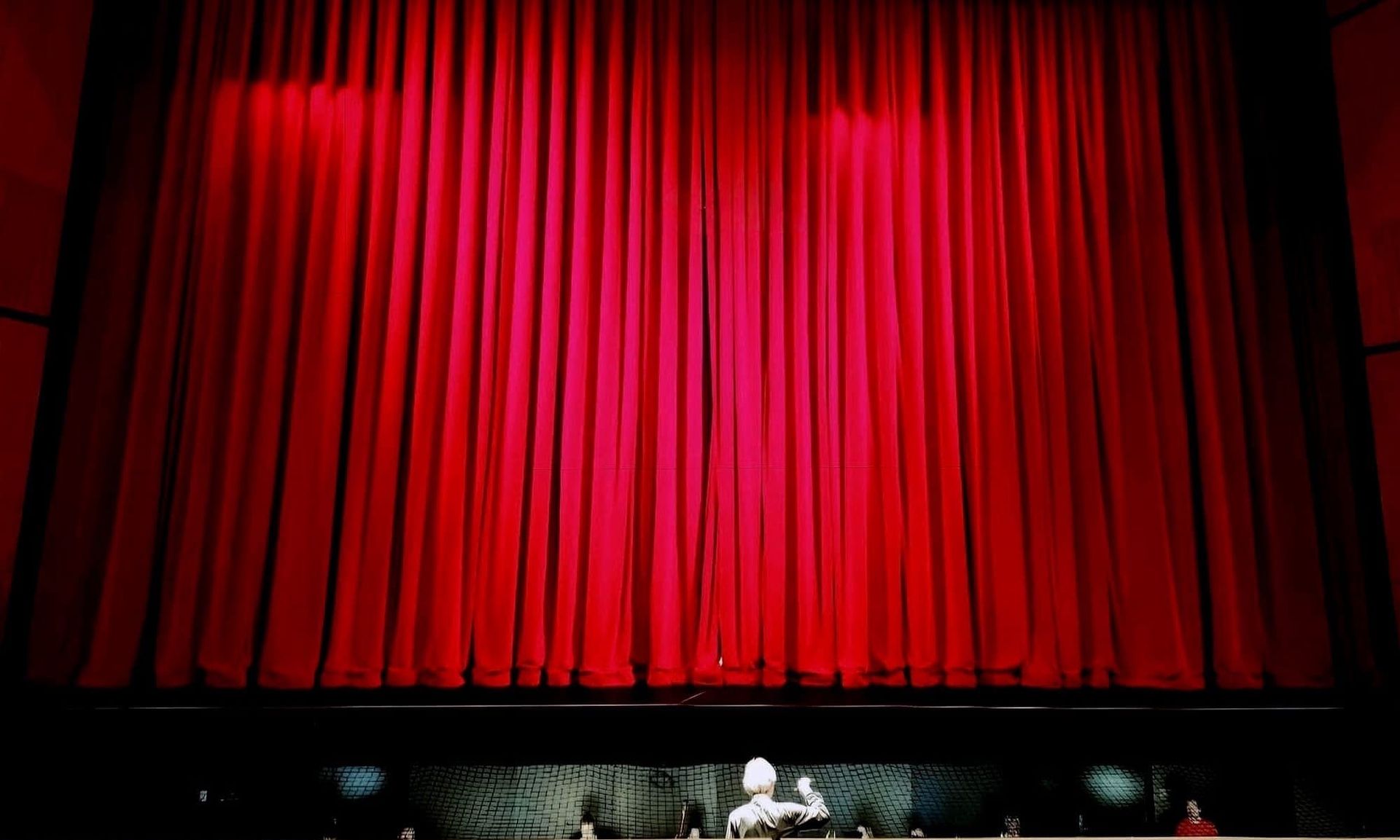 A conductor stands beneath a stage that is shrouded by a closed red curtain.