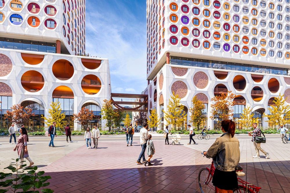 People stroll through a sunny courtyard at the base of two colourful highrise buildings.