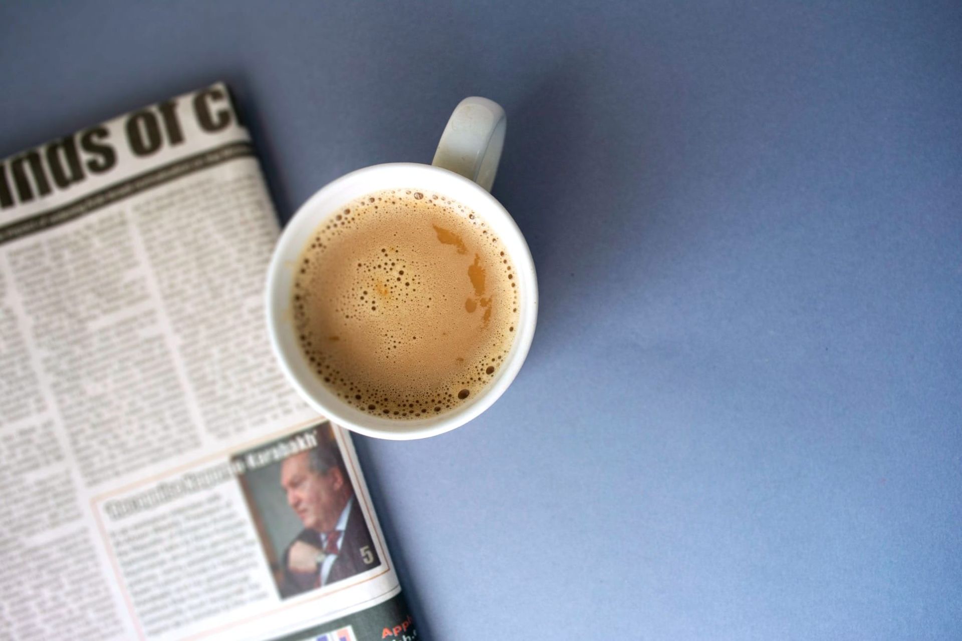 A cup of coffee sits beside a newspaper on a blue table.