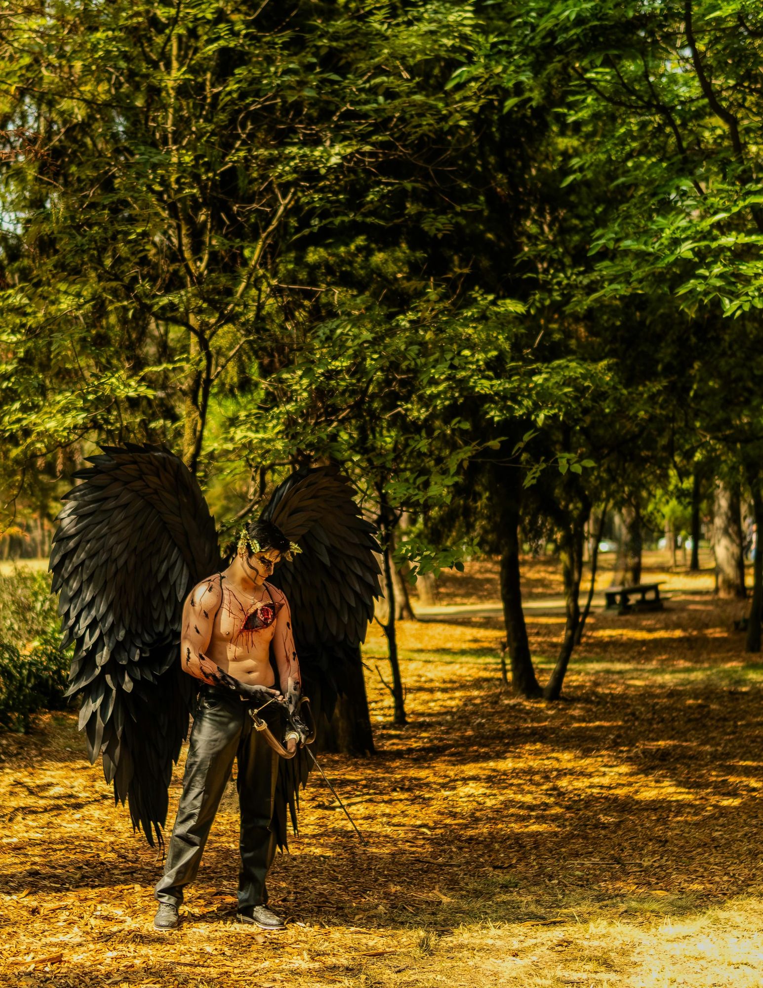 Man dressed in black feathered wings in front of an outdoor park of some kind. His skin is marked with fake blood