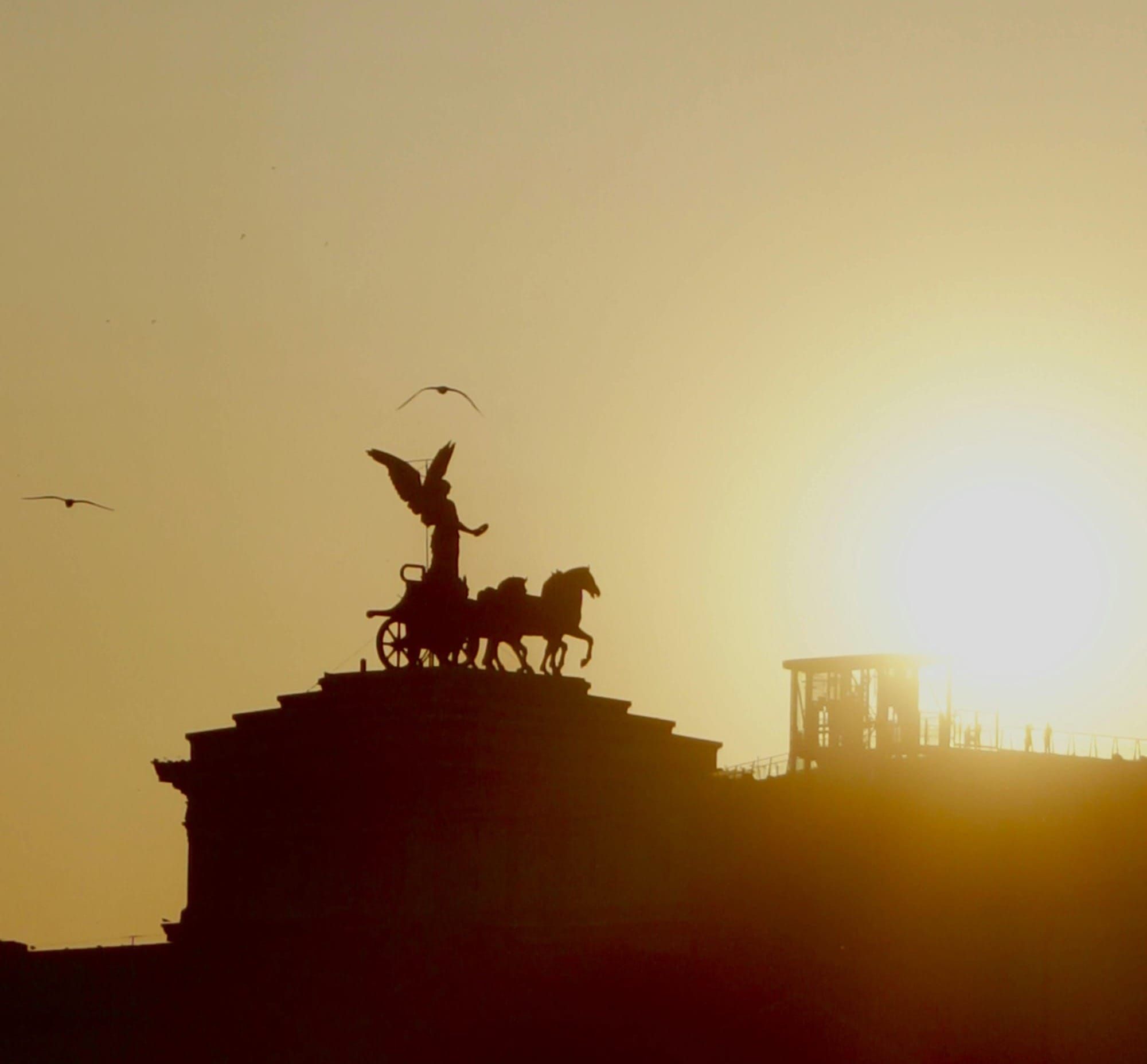 Silhouette of angel statue by a chariot of horses on a building in front of a large golden sun