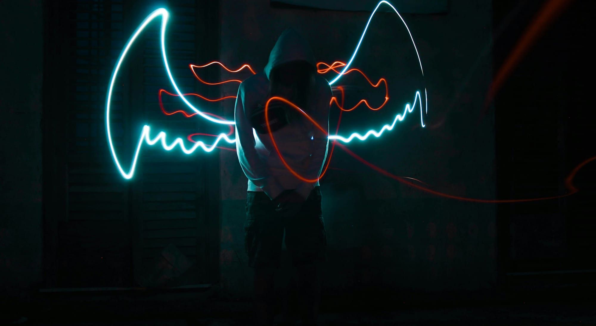 Dark image of someone in a white hoodie with their posture folded forward. Neon lights in the shape of wings and something red and ribbonlike emerge from their back