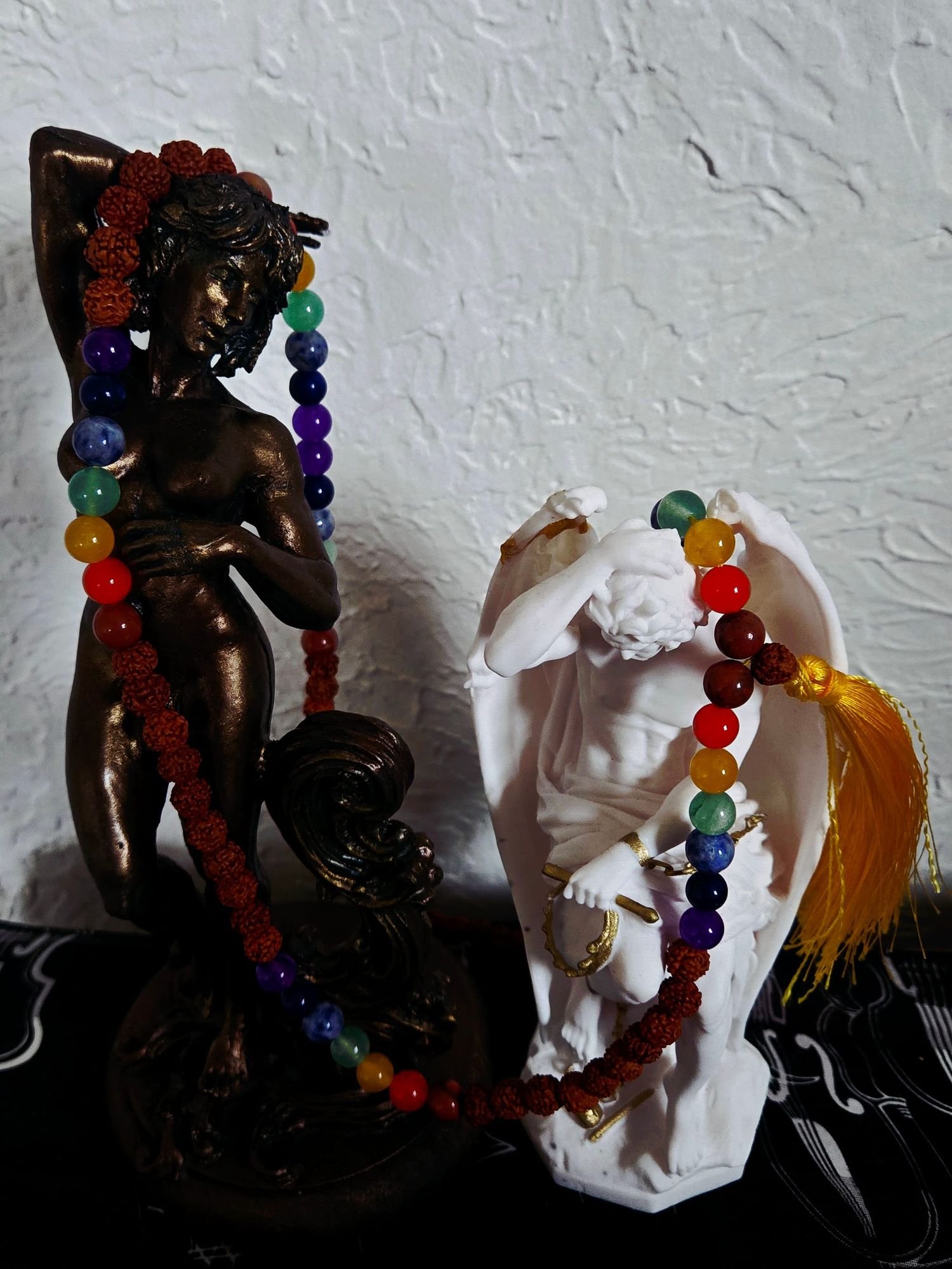 A bronze intersex statue stands next to a mostly white statue of genie du mal. They are wrapped together with mala beads, the guru bead and tassel laying across Lucifer's wing