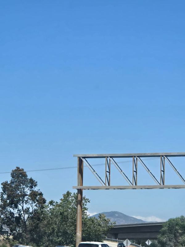 Photo taken from highway of a mountaintop with small clouds lining up just behind it. The front of it is cut off by an expressway, but then there's some nice green trees in front of that.