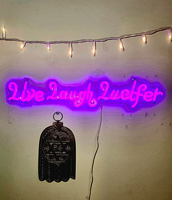 Purple neon sign declares "Live Laugh Lucifer." Hanging right under it by a nail is a metal hand of G-d with a large centered Star of David