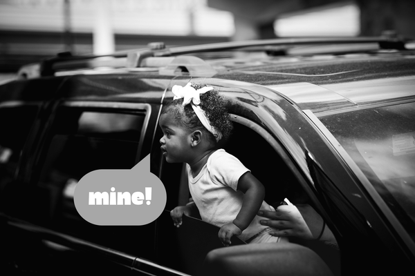 A Black toddler with curly hair and wearing a bow leans out of an SUV passenger window. A speech bubble underneath their mouth says MINE!