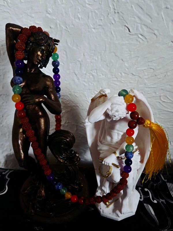 A bronze intersex statue stands next to a mostly white statue of genie du mal. They are wrapped together with mala beads, the guru bead and tassel laying across Lucifer's wing