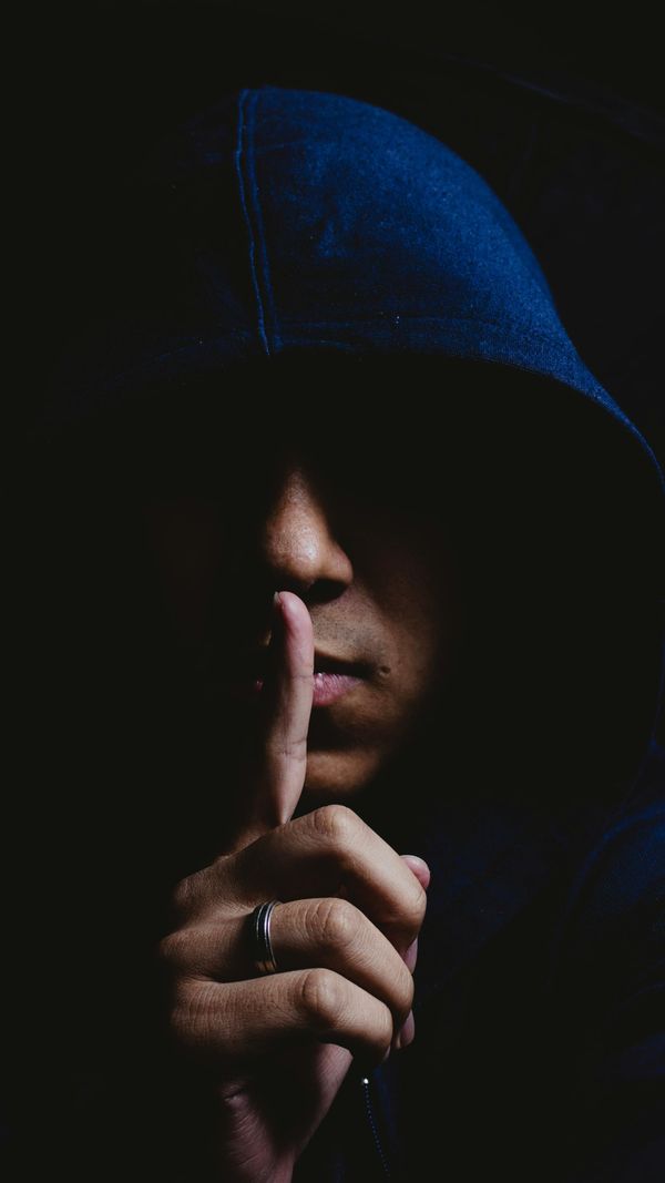 A male-presenting person mostly in shadow and wearing a dark blue hoodie which covers their eyes holds their index finger in front of their mouth in a shushing gesture