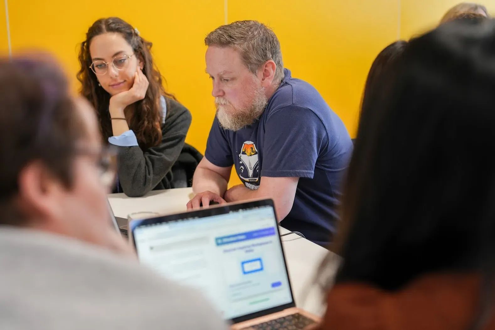 A group of four adults are working in pairs, looking at two laptops on a table.