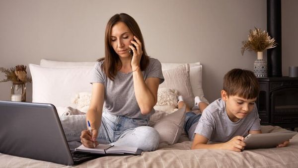 Mother talking on the phone, taking notes in a notebook. Her son is playing on an tablet next to her.