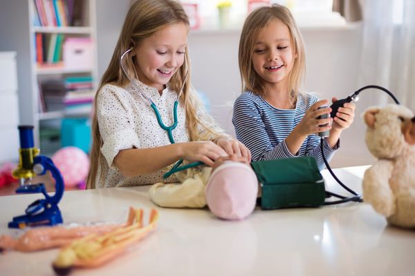 Two young girls smiling while role playing as doctors. They're using a stethoscope and a manual blood pressure monitor on a baby doll. 