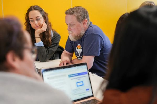 A group of four adults are working in pairs, looking at two laptops on a table.