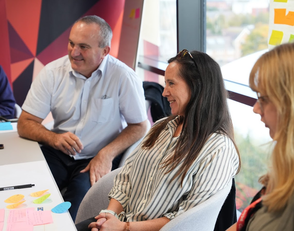 Man and woman smiling at someone out of frame. In front of them is an a3 piece of paper covered in sticky notes. 