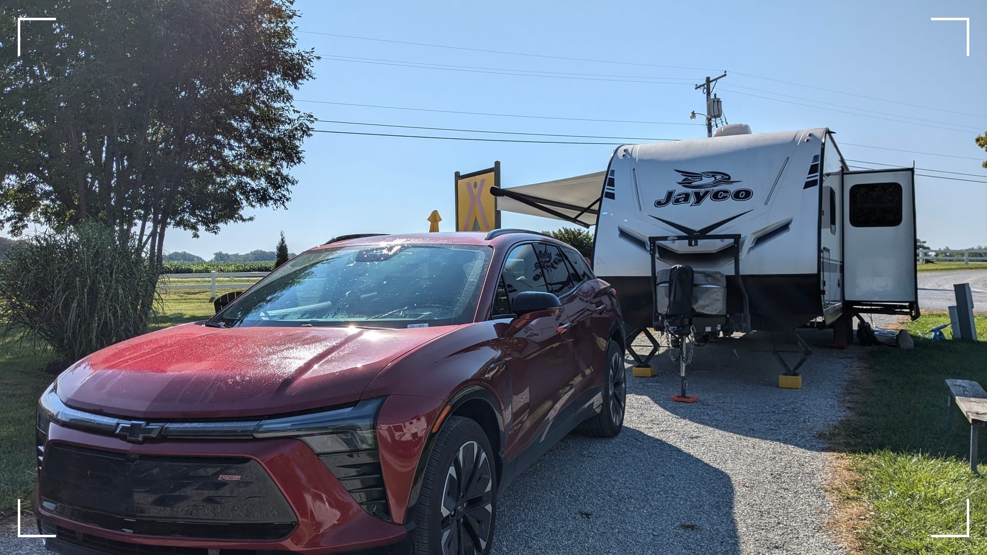Travel trailer set up for camping with a red electric vehicle in the front at a campground with a farm field in the background.