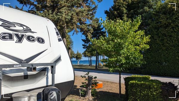 A Jayco travel trailer parked in a campground with trees in the background. Beyond the trees, you can see Lake Huron and the Mackinac Bridge. 