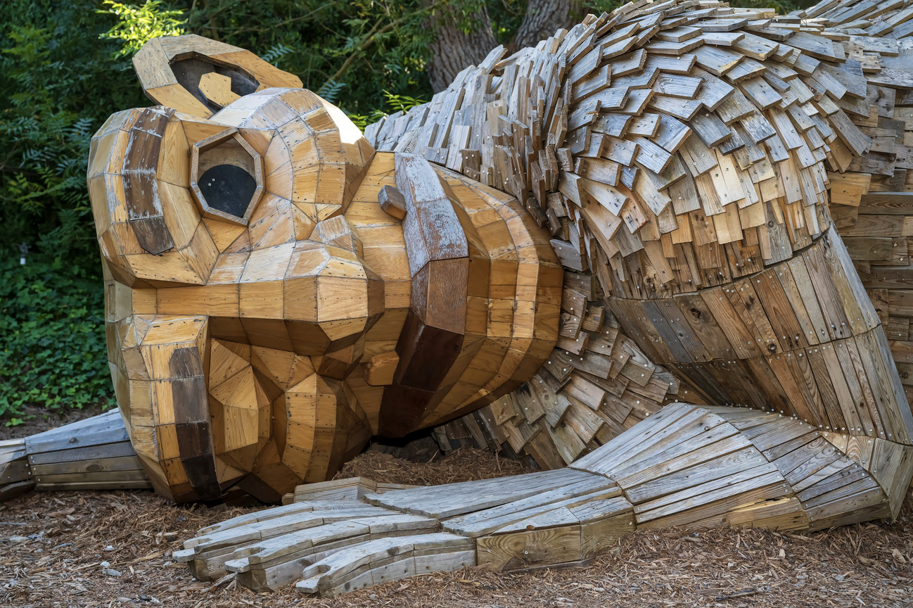A large wooden sculpture shows a troll laying on the ground listening to the earth with one eye shut.