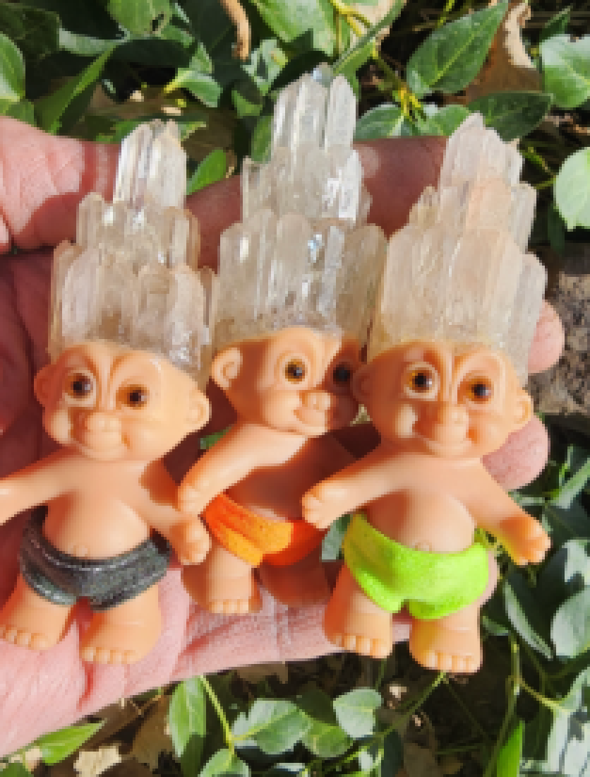 Three tiny plastic troll dolls with wide brown eyes and wearing different colored shorts. Their hair is made of quartz crystal.