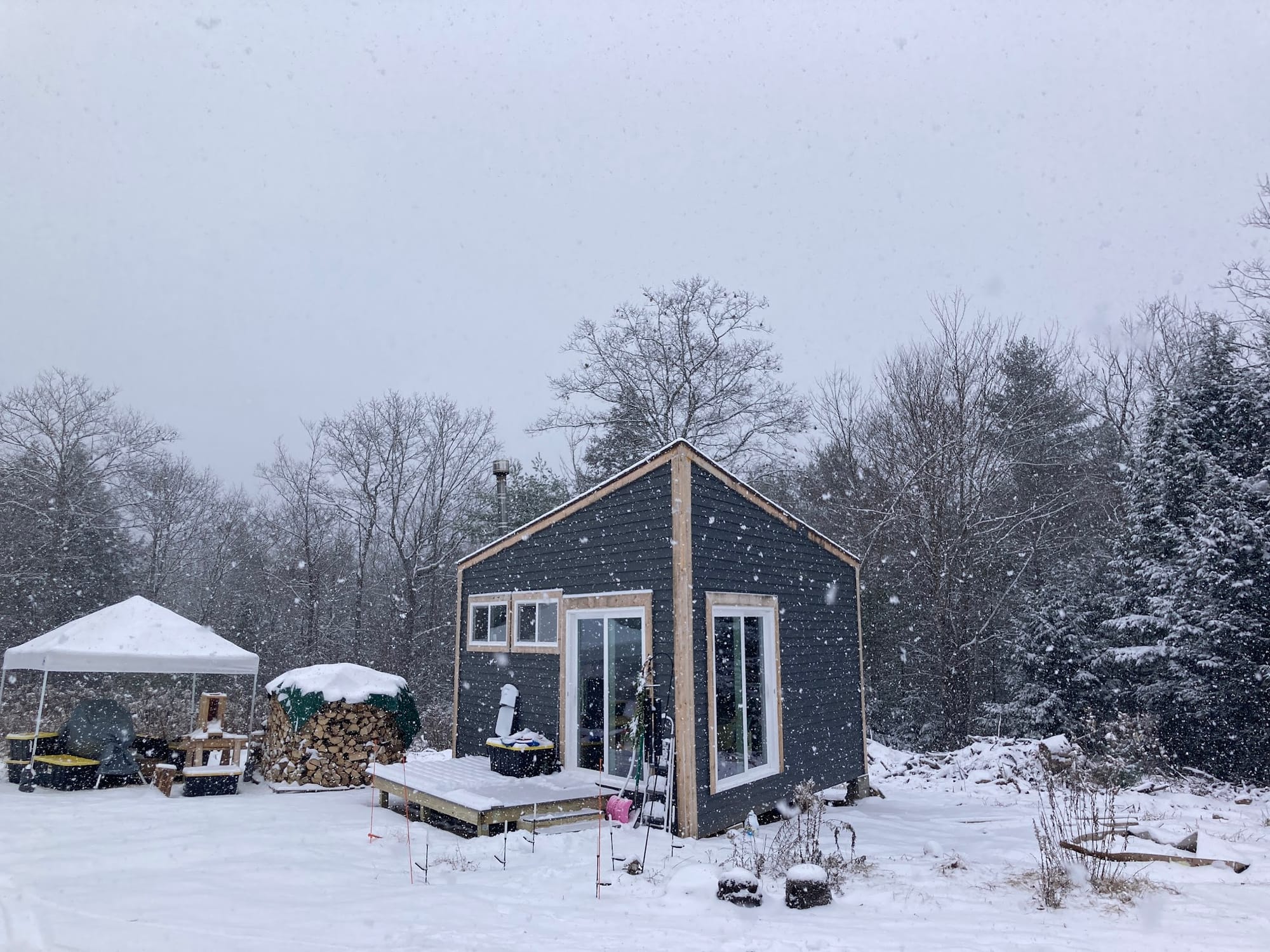 A small grey house with large glass doors and a deck sits amidst heavily falling snow.