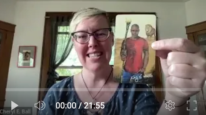 White woman with short blonde hair and glasses smiles and holds up a tarot card, the King of Wands