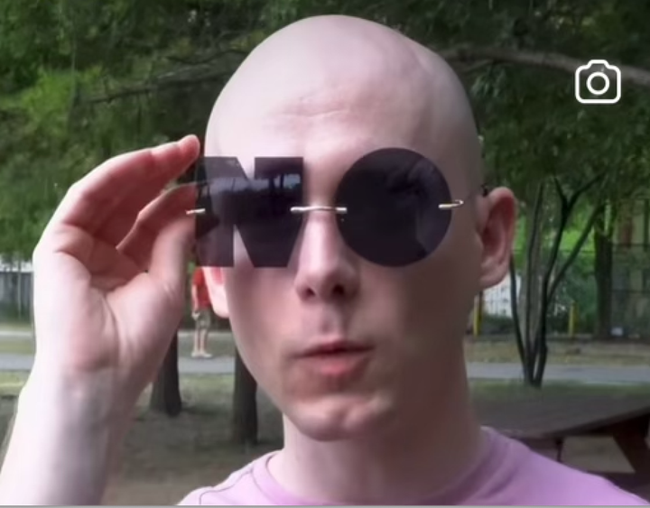 Bald person with a pink tee shirt holding sunglasses on their face. The lenses read NO.