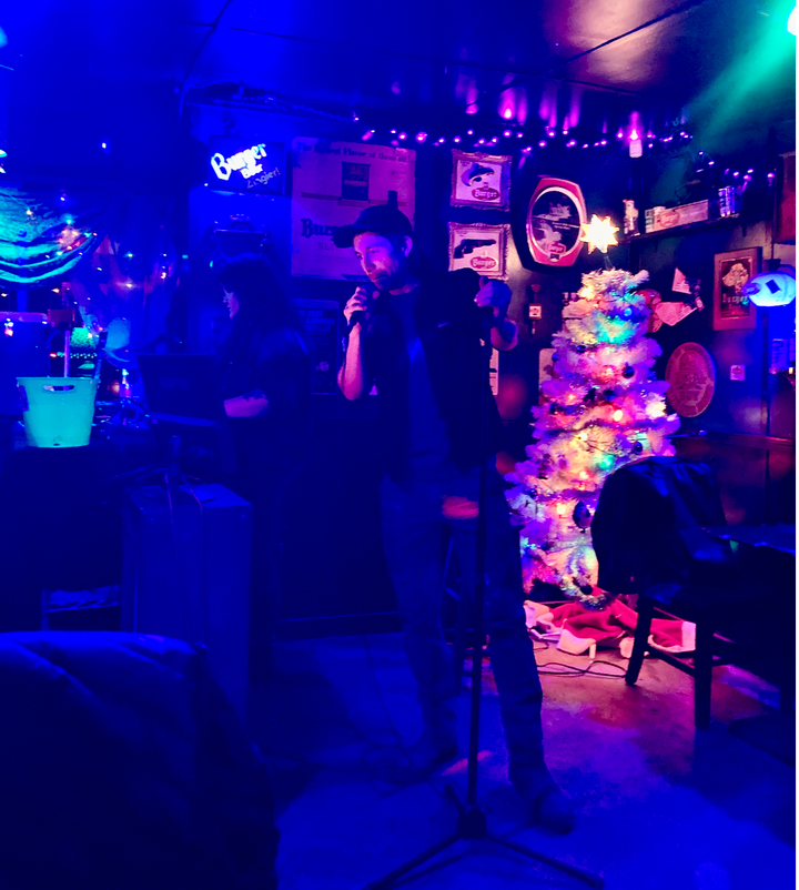 A guy in a hat stands in a dark bar behind a mic. Behind him are a white, lit Christmas tree and a female DJ